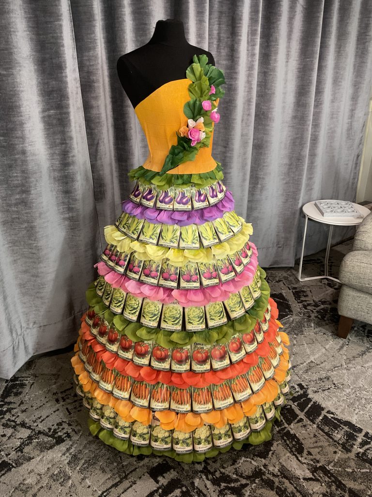 A mannequin dressed in a floor-length gown handcrafted from hundreds of layered vegetable seed packets arranged in rainbow-colored tiers, with green leaf accents and floral details at the shoulder, created by Retailworks for a spring window display at Diedrich Jewelry.
