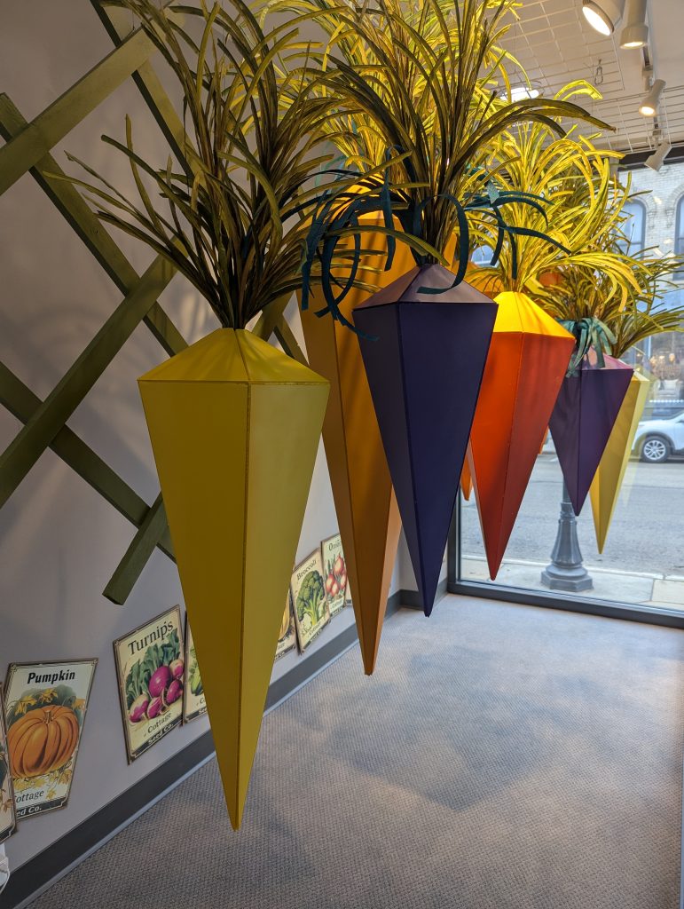 A spring retail window display created by Retailworks for Diedrich Jewelry, featuring large handcrafted paper carrot sculptures in yellow, orange, red, and purple hanging above colorful vegetable seed packet arrangements, set against a backdrop of green lattice and tropical foliage.