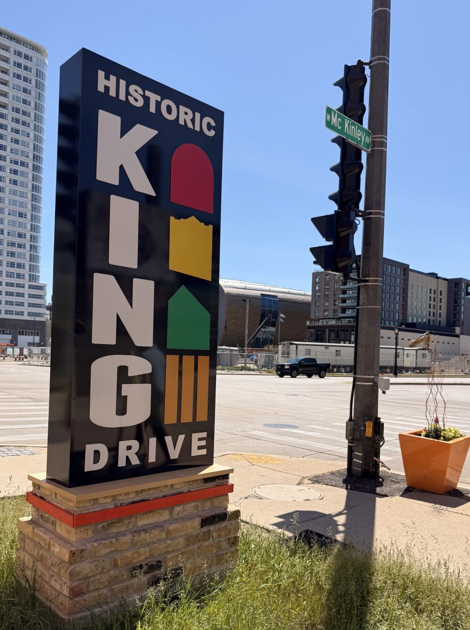Historic King Drive sign at West McKinley Avenue in Milwaukee, Wisconsin, with downtown buildings and urban streetscape under a clear blue sky.