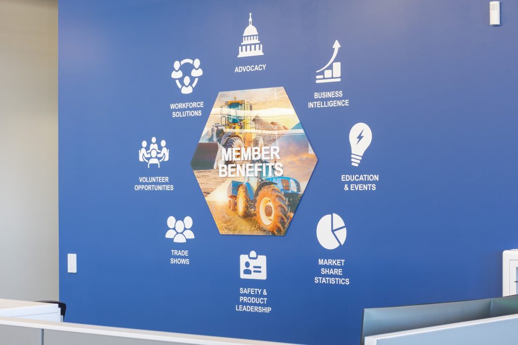 AEM office wall display on a deep blue painted wall featuring a central hexagonal printed graphic of construction and agriculture equipment labeled "Member Benefits," surrounded by white vinyl icons and labels highlighting seven member benefits including Advocacy, Business Intelligence, Education & Events, Market Share Statistics, Safety & Product Leadership, Trade Shows, and Workforce Solutions.