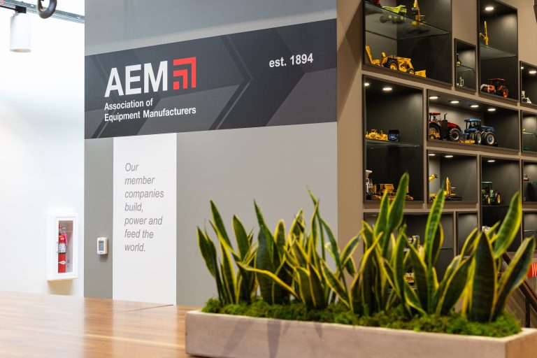 AEM headquarters lobby display featuring a branded wall graphic with logo, "est. 1894," and the mission statement "Our member companies build, power and feed the world," alongside a lit display case showcasing scale models of construction and agriculture equipment.