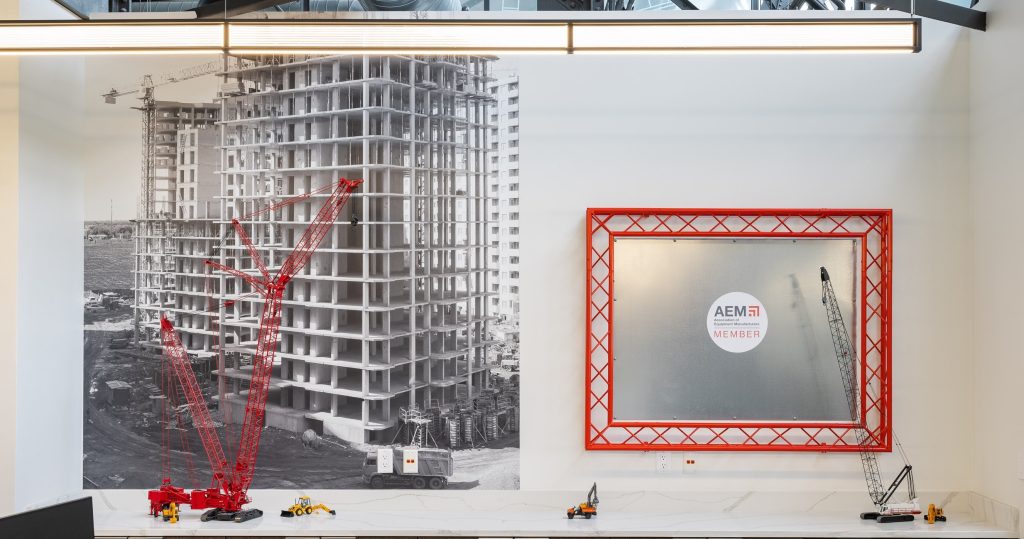 AEM office display featuring a large black and white construction photo mural with red crane accents, alongside a bold red metal lattice frame enclosing a mirrored panel with the AEM Member logo, with scale model construction equipment arranged on a white shelf below.