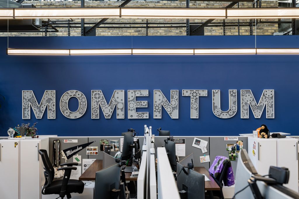 Large dimensional "MOMENTUM" sign mounted on a deep blue wall above an open office workspace, with each letter featuring CNC-cut gear and cog details, set against the exposed brick and industrial ceiling of a building.