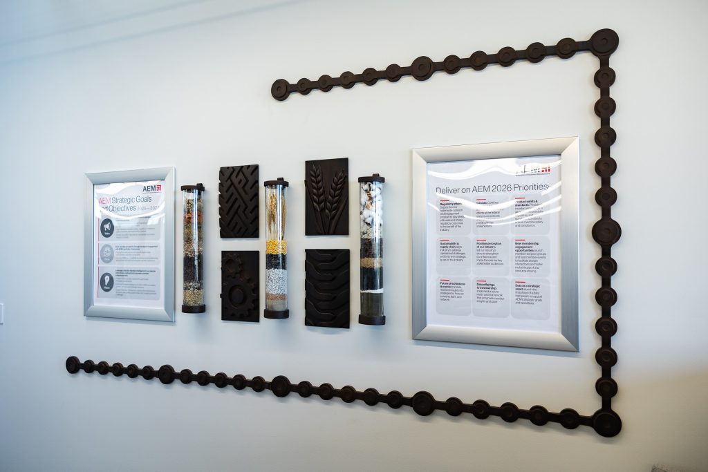 AEM office wall display featuring two aluminum-framed informational panels showing AEM Strategic Goals and 2026 Priorities, flanking a series of CNC-cut dark relief panels depicting tire treads and a wheat stalk, with clear tubes filled with agricultural materials, all connected by a decorative dimensional roller chain border along the top and bottom of the display.