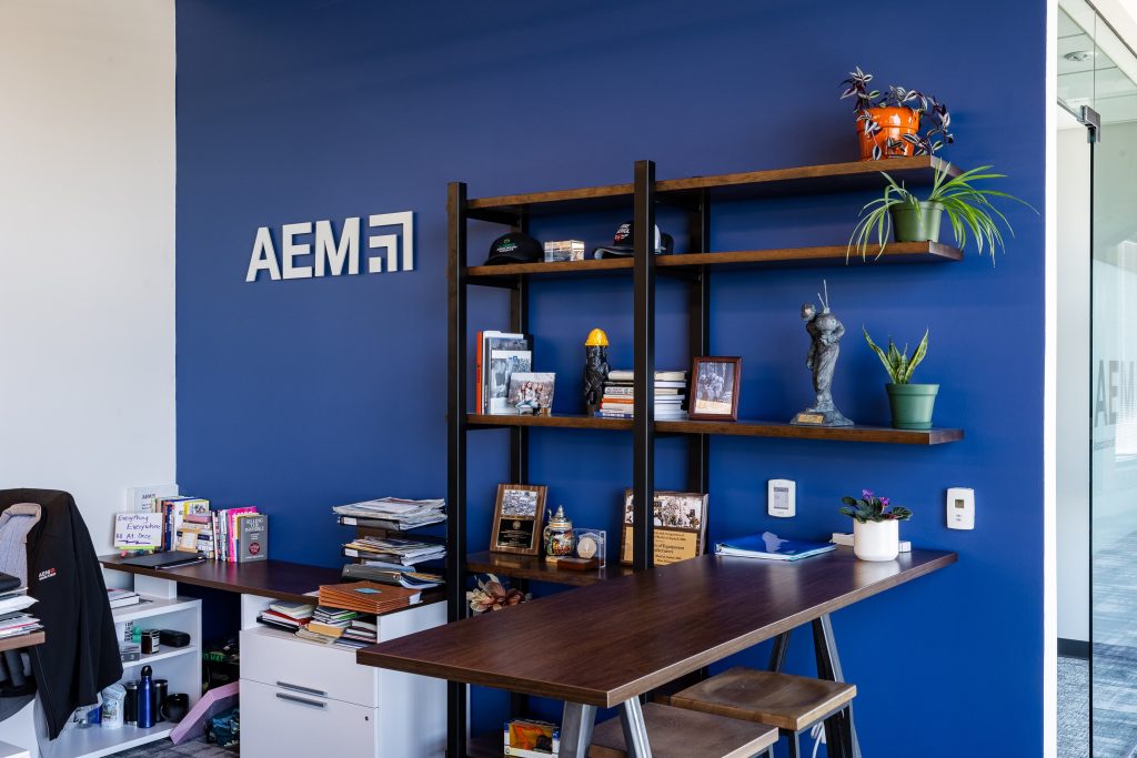 AEM office display featuring a large black and white construction photo mural with red crane accents, alongside a bold red metal lattice frame enclosing a mirrored panel with the AEM Member logo, with scale model construction equipment arranged on a white shelf below.