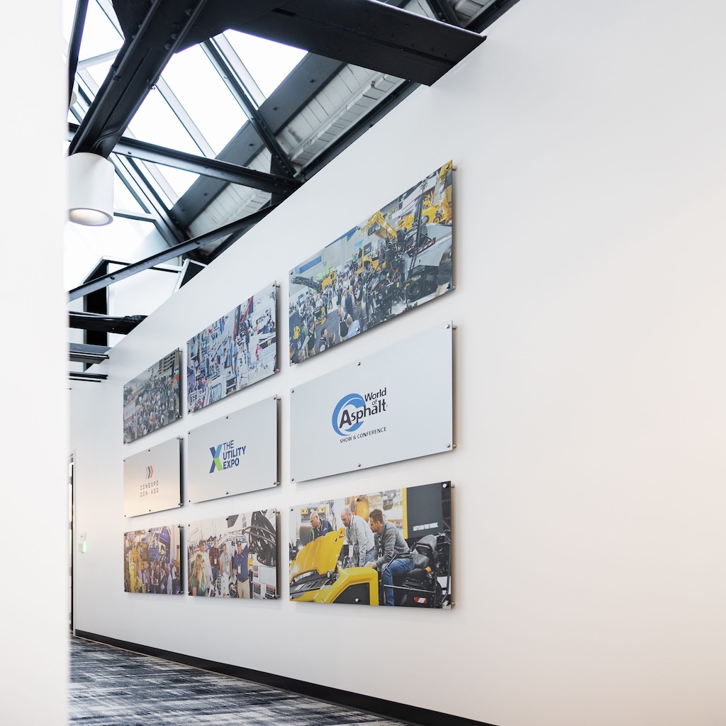 Hallway wall display at AEM headquarters featuring a grid of printed aluminum photo panels showing industry trade shows alongside printed acrylic panels mounted with standoffs displaying trade show and expo logos.