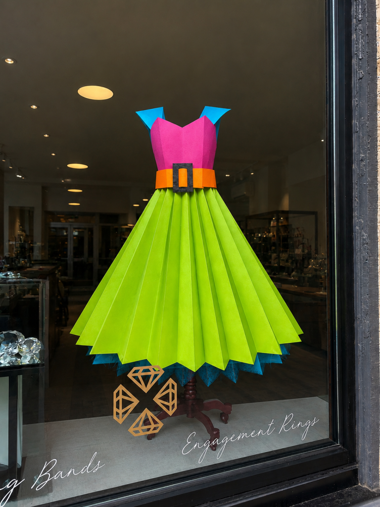 Colorful paper dress display in a jewelry store window, featuring a bright pink and blue bodice, neon green pleated skirt, orange belt, and gold diamond graphics.